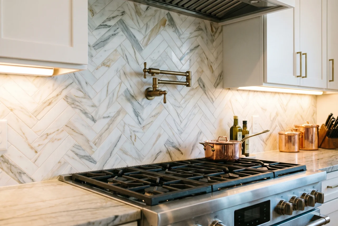 Marble herringbone tile backsplash with brass pot filler over a stainless gas range